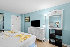 A hotel room with a bed, a television, a dresser, a mirror, and a lamp. - Deluxe Oceanfront Suite | King Bed (Myrtle Beach)