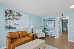 A living room with a brown leather sofa, a white armchair, a small table, and a lamp. There are two framed pictures on the wall. - Deluxe Oceanfront Suite | King Bed (Myrtle Beach)