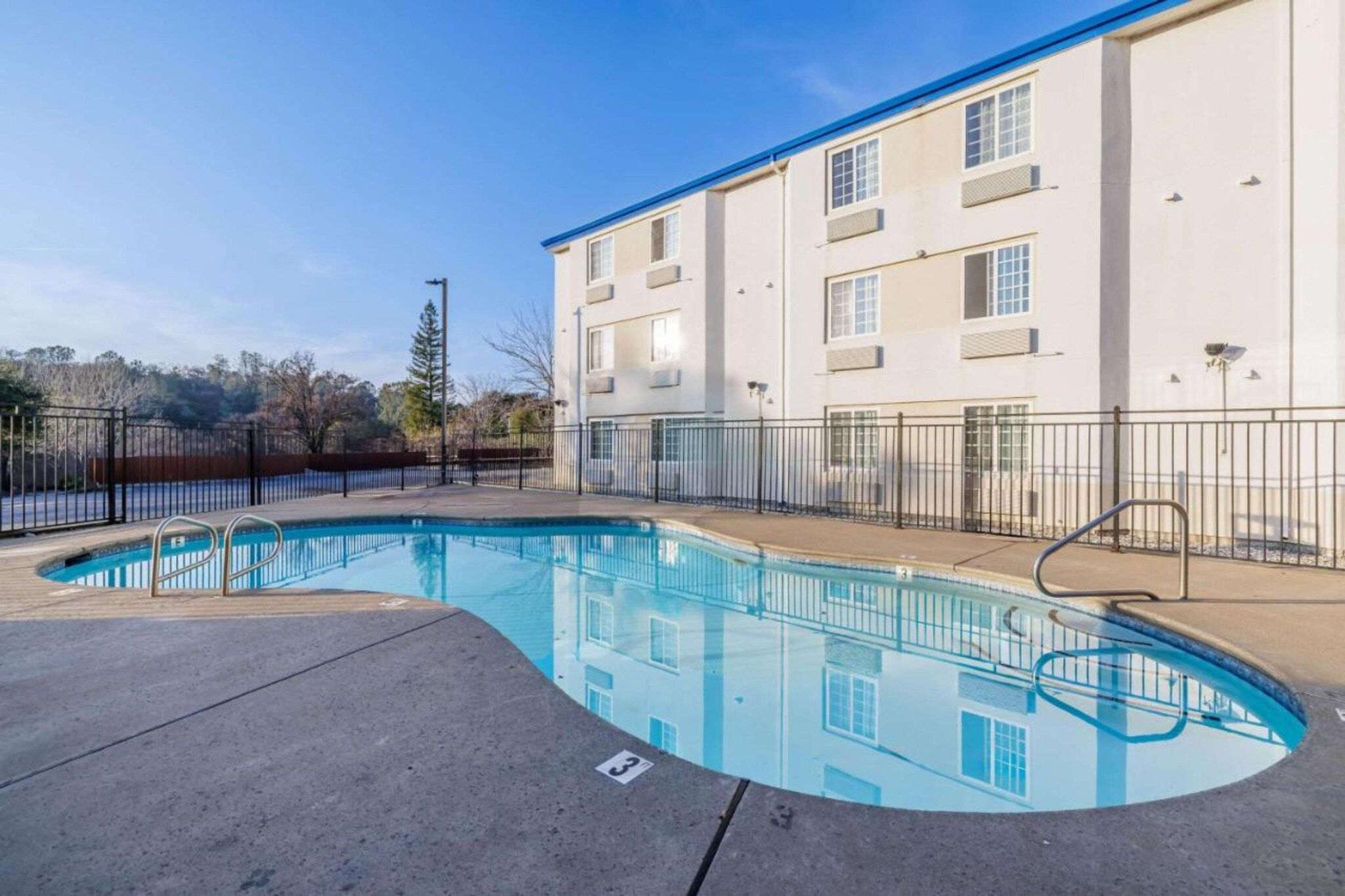 Una piscina de borde curvo, rodeada por una cerca metálica, junto a un edificio de varios pisos con ventanas.