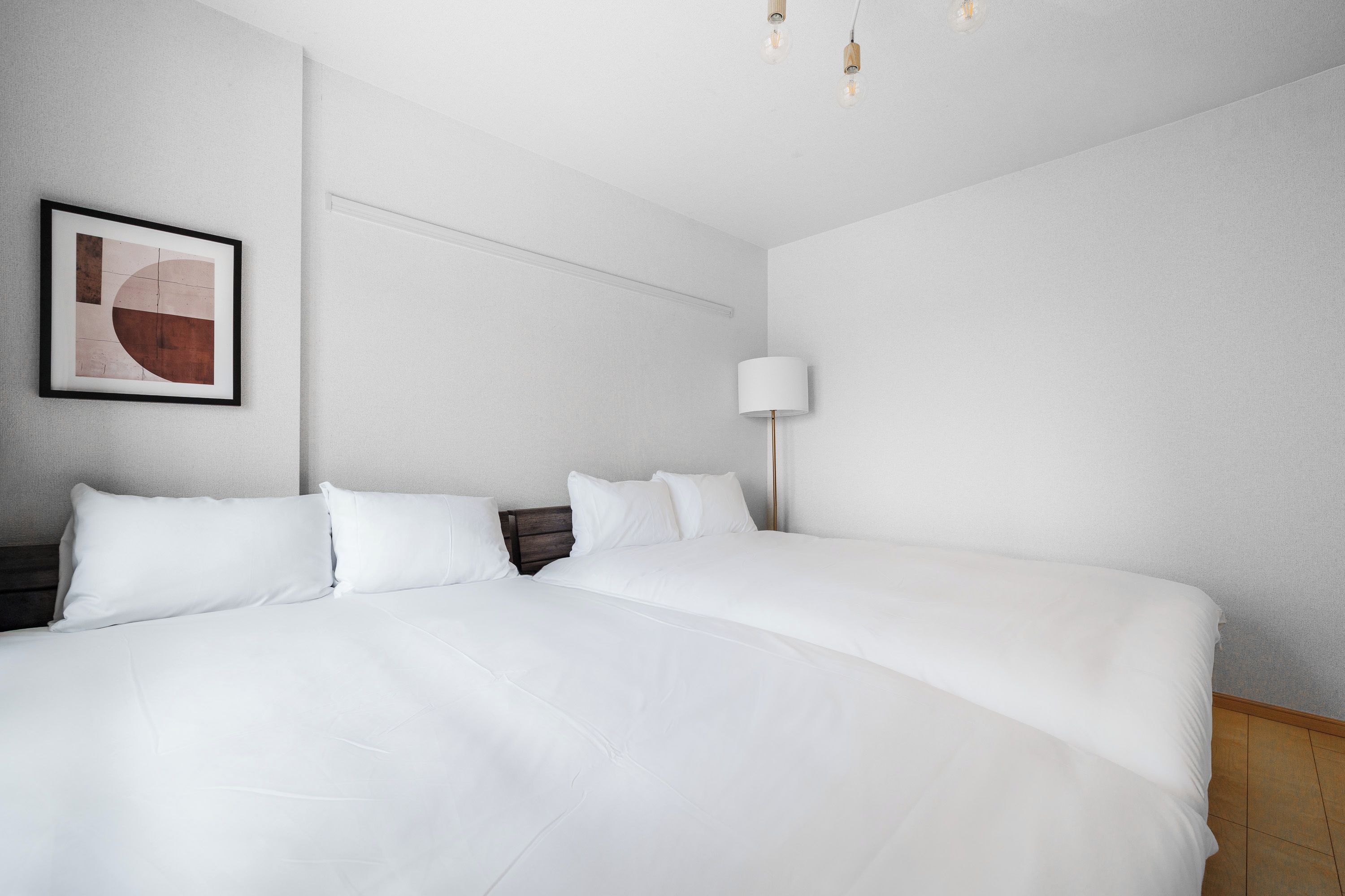 Una cama bien hecha con ropa de cama blanca y un cabecero, una lámpara de pared y una obra de arte abstracta enmarcada.