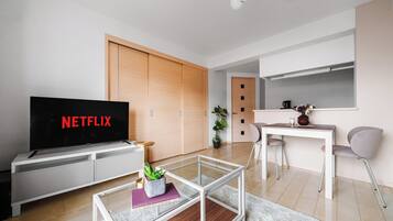 Una sala de estar moderna con un televisor de pantalla plana mostrando 'NETFLIX', una mesa de centro de vidrio, una planta en maceta y una zona de comedor con una mesa y sillas blancas.