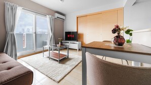 A modern living room with a glass coffee table, a sofa, a dining table with chairs, a TV, and a city view through large windows. - E-US Naebo Station 407 (Sapporo)
