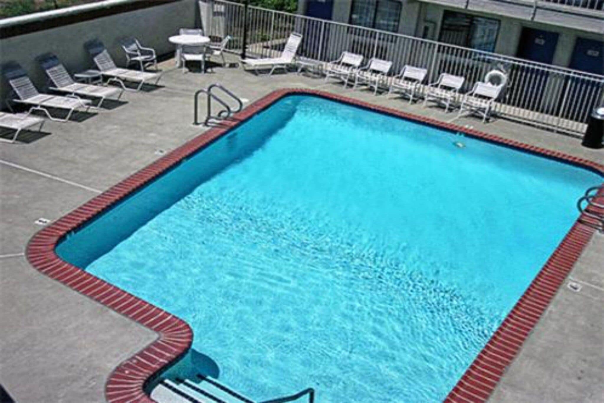 Piscina retangular com borda vermelha, rodeada por espreguiçadeiras brancas e uma pequena mesa.