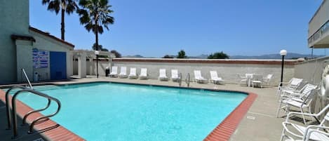 A rectangular swimming pool with a red brick border, surrounded by white lounge chairs, palm trees, and a clear sky.