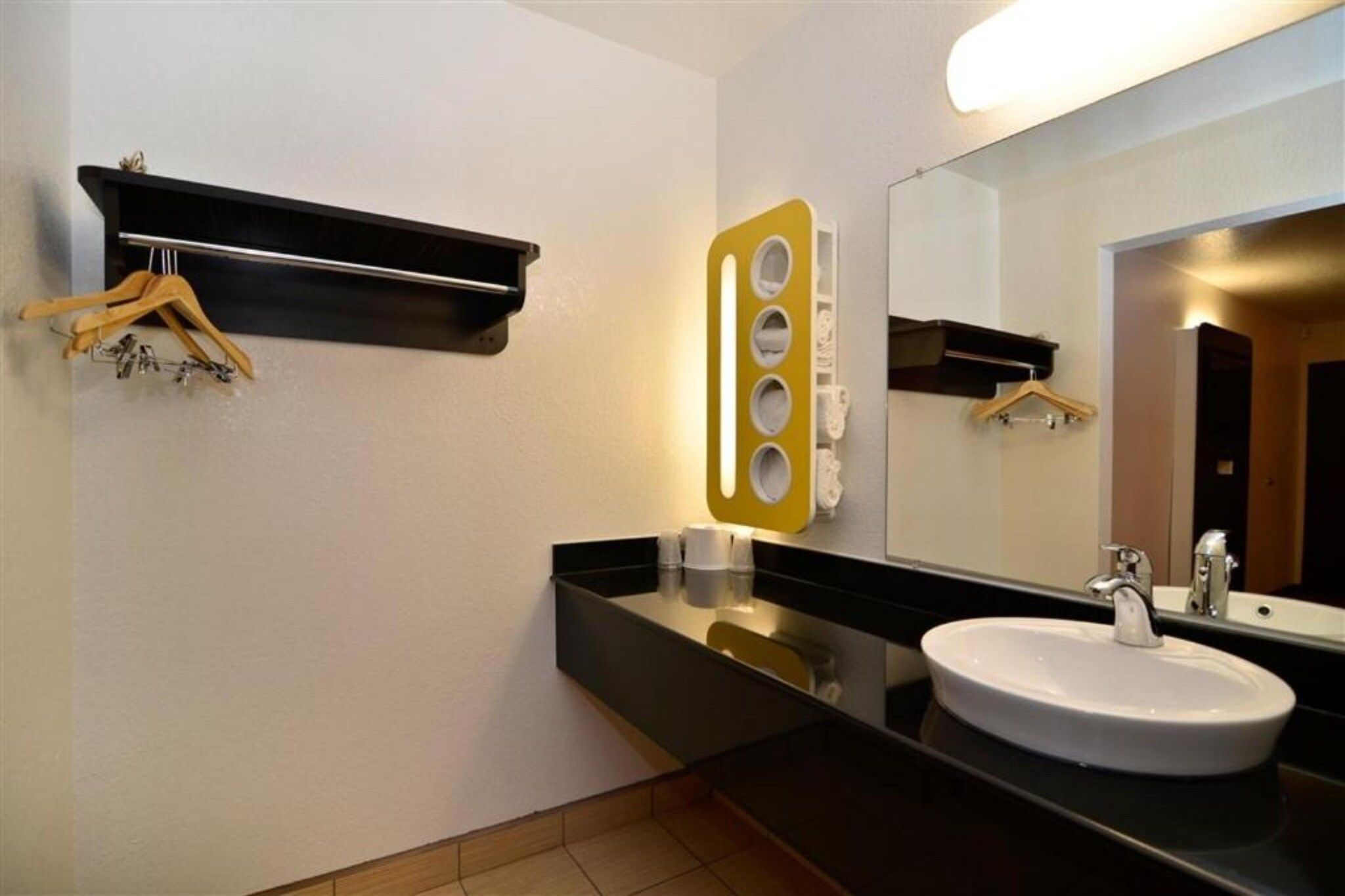 Un baño con encimera negra, lavabo blanco, espejo y estante con perchas.