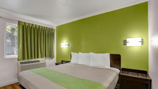 A hotel room with a bed, a nightstand, a wall-mounted lamp, a window with curtains, and an air conditioning unit. - MS6 King City | 1 Queen Bed | NS MicFridge (King city)