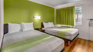 Una habitación de hotel con dos camas, una pared verde, un ventanal con cortinas y una mesita de noche con lámpara.
