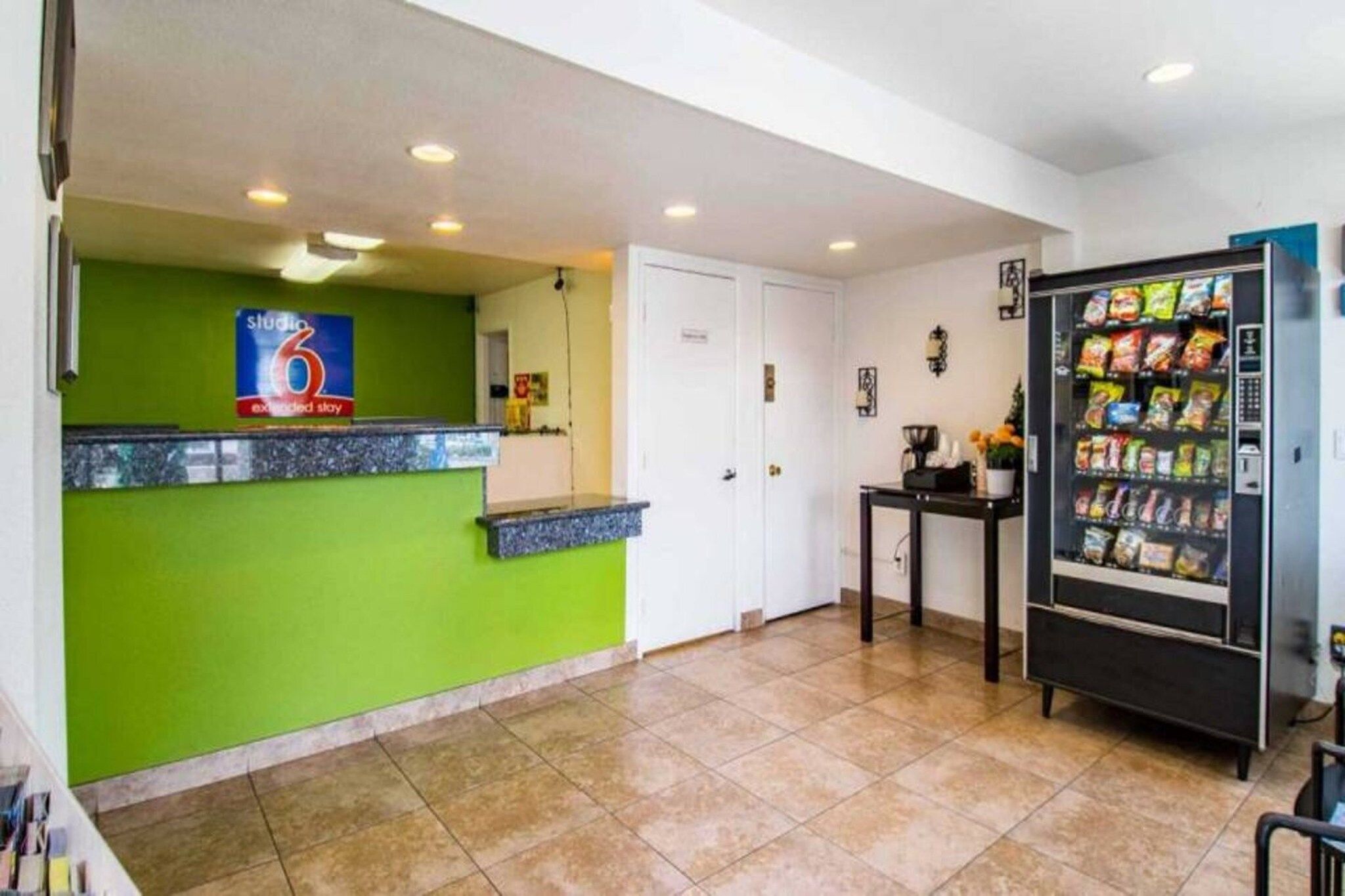 A reception area with a green wall, a counter, a door, a small table with a vase, and a snack vending machine.
