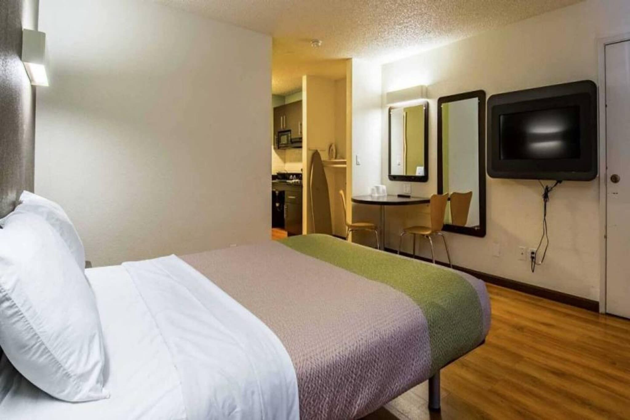 Une chambre d’hôtel avec un lit, un bureau, une chaise, une télévision et, au fond, une petite cuisine.