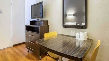 A hotel room with a wooden table, a television on a stand, a mirror on the wall, and a chair.