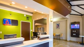 Zona de recepción de hotel con una pared verde, mostrador de recepción, televisor y un área de estar con sillas.