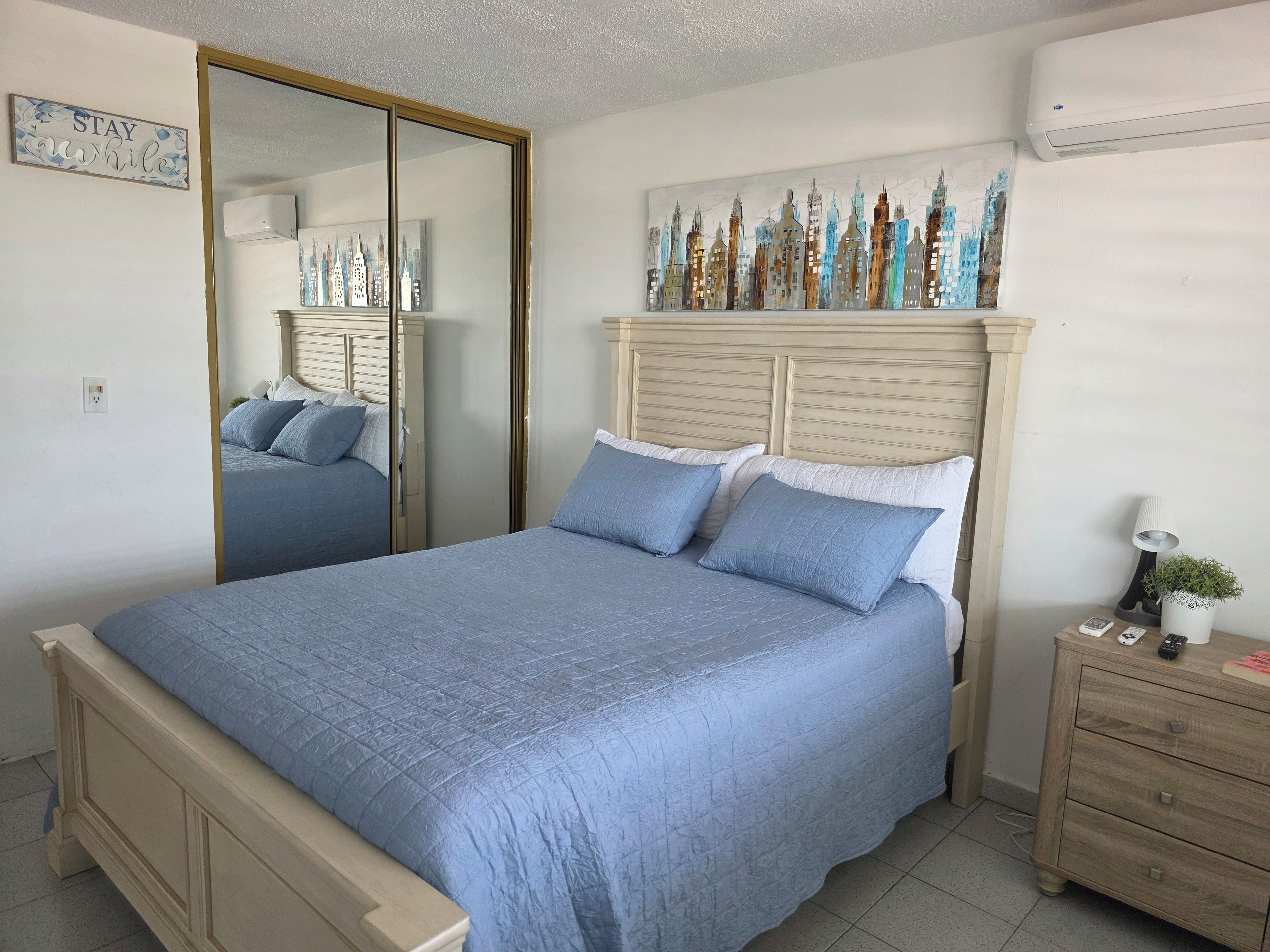 A bedroom with a bed, a bedside table, a mirror, and a wall-mounted air conditioner.