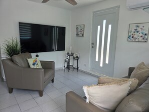 Ein Wohnzimmer mit einer Couch, einem Fernseher, einer Tür mit vertikalen Jalousien und einer Pflanze.