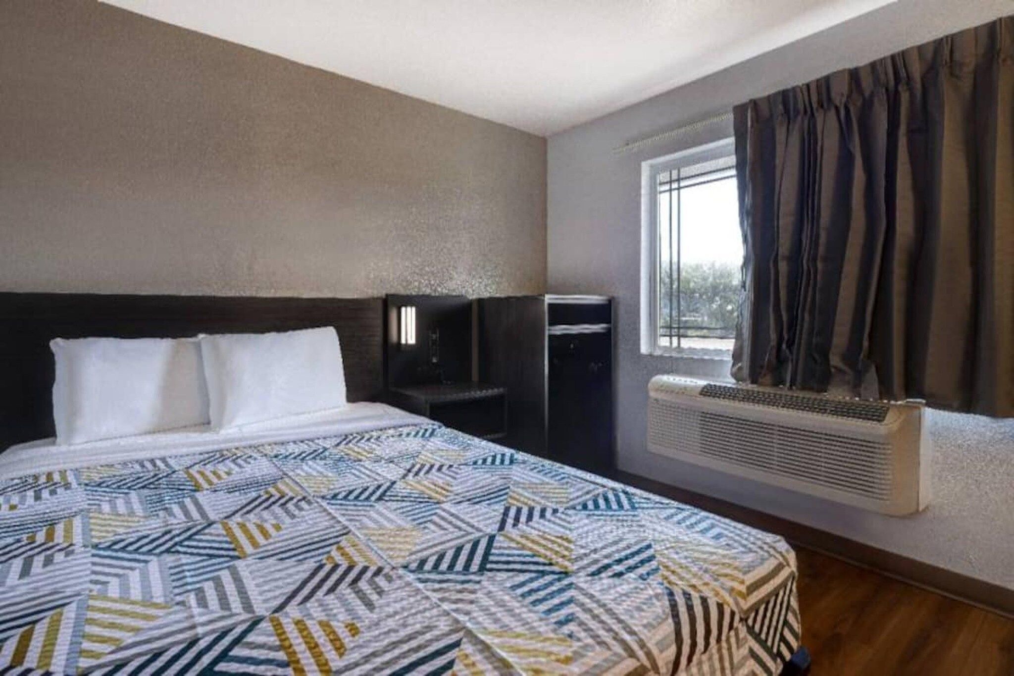 A hotel room with a bed, a heater, a window with curtains, and a wall-mounted air conditioning unit.