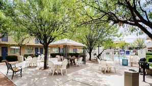Un patio al aire libre con mesas y sillas, rodeado de árboles y un edificio con puerta azul.