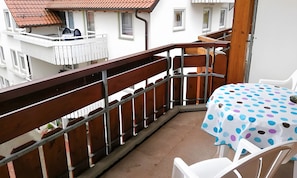 Un balcon avec une balustrade en bois, une table recouverte d’une nappe à pois et des chaises en plastique blanches.