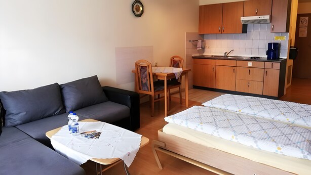 Ett kompakt vardagsrum med en soffa, ett litet bord med en vattenflaska, en trästol, en säng med ett mönstrat överkast och ett litet kök med träskåp och en kakelbeklädd vägg bakom diskbänken.