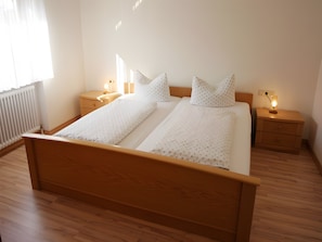 Una cama doble con estructura de madera, dos mesitas de noche con lámparas, un radiador y una ventana con cortinas.