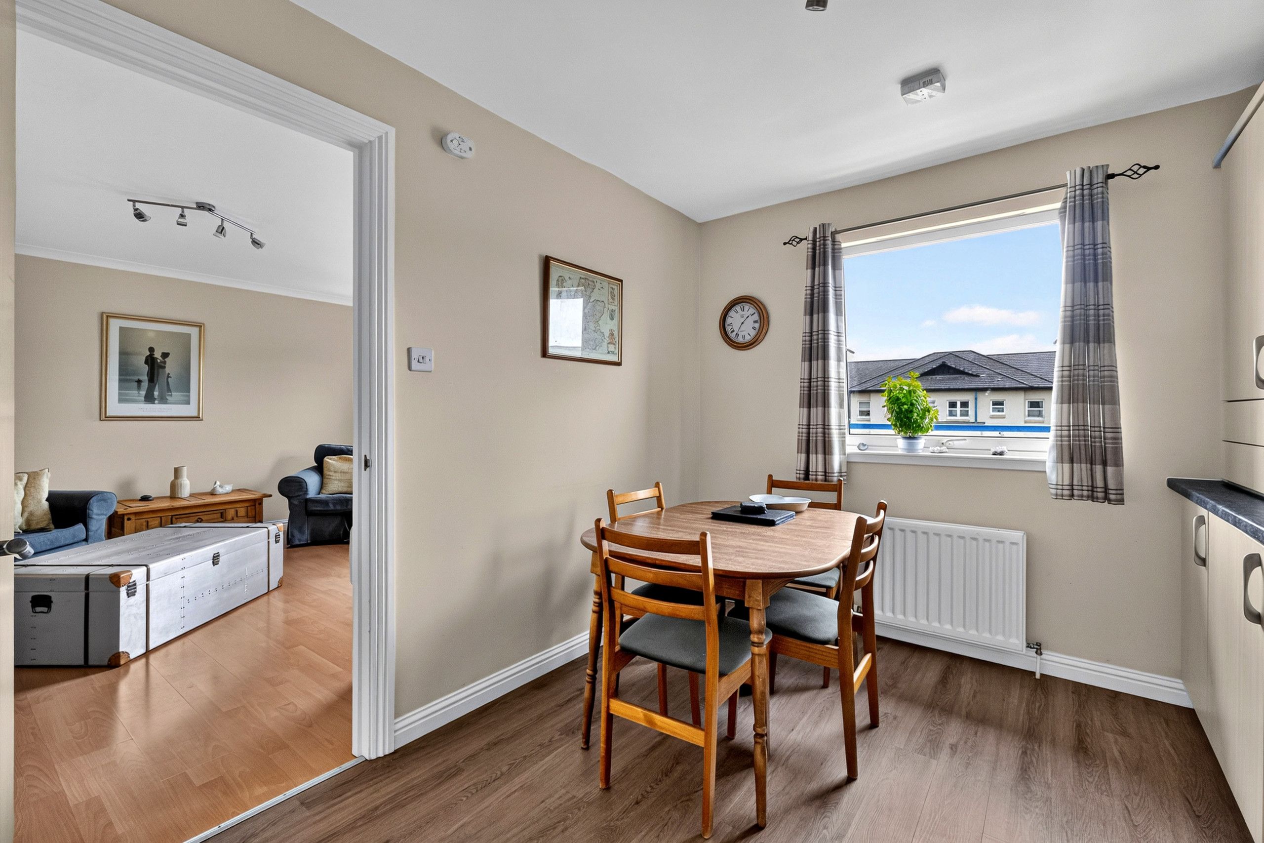 A dining area with a wooden table and chairs, a window with curtains, a clock, and a radiator.