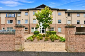 A multi-story residential building with a central entrance, a small garden area with shrubs and a tree, and a paved walkway leading to the entrance. - Ness-side Apartment (Inverness)
