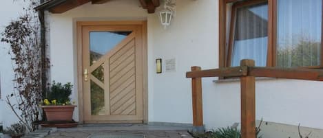 Entrada de un edificio con puerta de madera, planta en maceta, sendero y ventana con cortina.