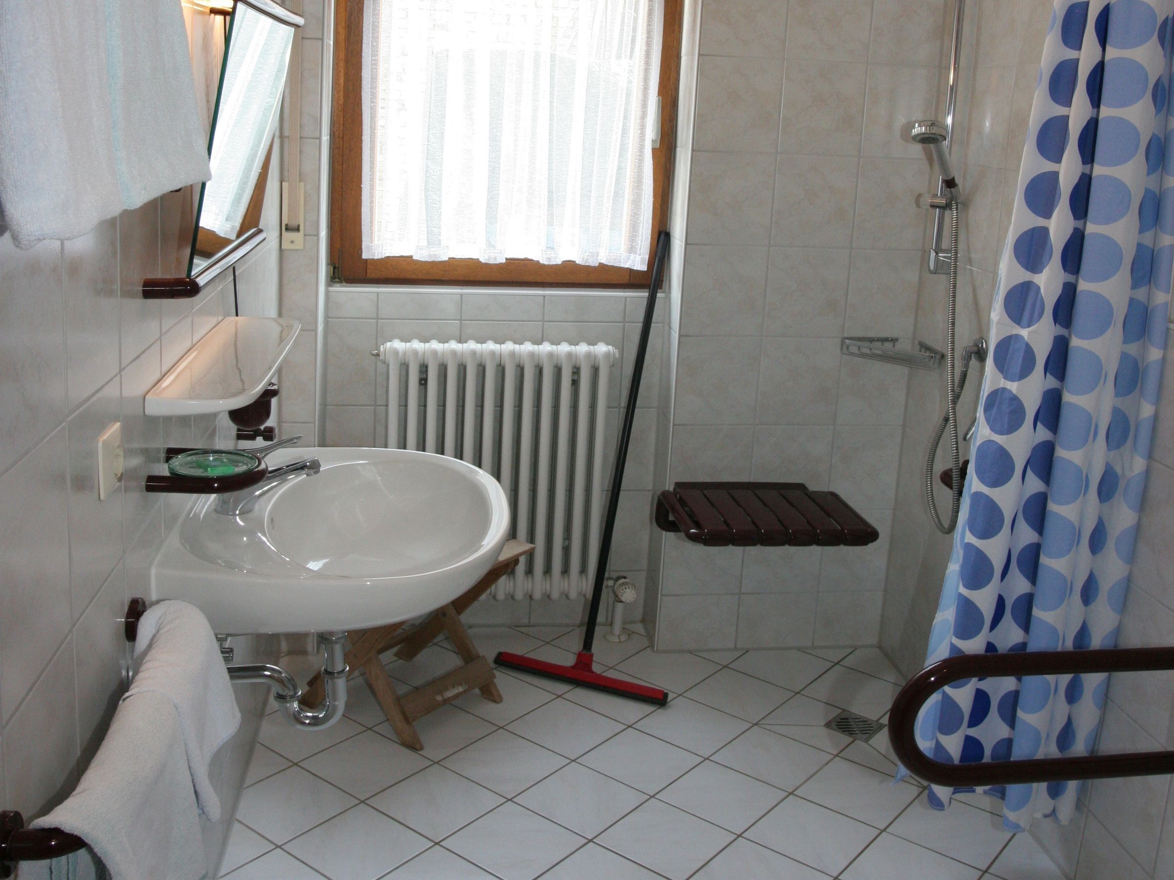 A bathroom with a white sink, a shower, a window with curtains, a radiator, and a wooden step.
