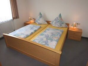 Una cama doble con ropa de cama floral, dos mesitas de noche con lámparas, un radiador y una ventana con cortinas.