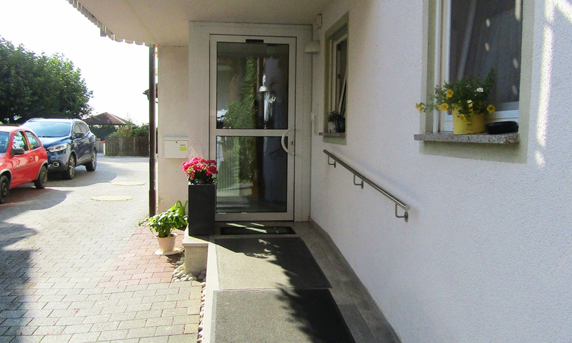 Une entrée d’immeuble avec une porte vitrée, un petit espace extérieur aménagé avec des plantes en pot et une fenêtre agrémentée d’une jardinière.