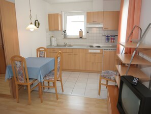 Cocina compacta con armarios de madera, una mesa de comedor con sillas, horno empotrado y televisor.