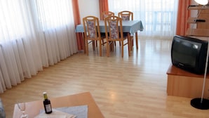 Uma área de refeições com mesa posta para dois, uma televisão sobre um suporte e uma janela com cortinas.