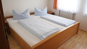 Cama de casal com roupa de cama branca e duas almofadas, cabeceira de madeira, mesa de cabeceira e janela com cortinas.