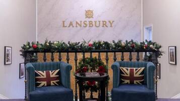 Ett rum med två blå fåtöljer, ett litet bord och en öppen spis som är dekorerad för jul. På väggen står ordet ”LANSBURY” med guldfärgade bokstäver.
