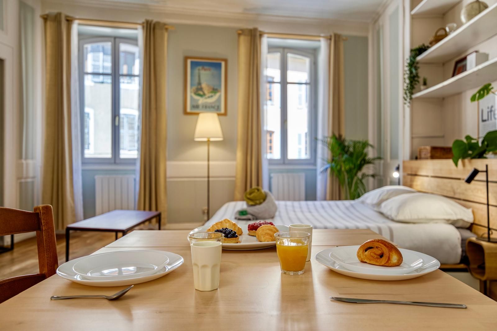 Ein Hotelzimmer mit Frühstückstisch, gedeckt mit Croissants, Obst und Saft, einem Bett mit weißer Bettwäsche und Blick auf die Stadt durch große Fenster.