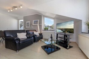 Een moderne zithoek met een leren bank in het zwart, een glazen salontafel en een flatscreen televisie aan de muur.
