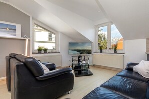 A modern living room with a black leather sofa, a glass coffee table, and a flat-screen TV mounted on the wall. - Holiday apartment with 1 bedroom (Ostseebad Binz)