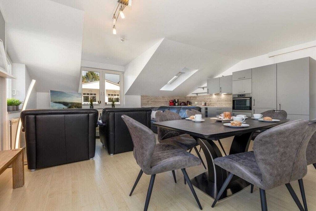 A modern kitchen with a dining area, featuring a high ceiling with skylights, dark grey upholstered seating, and a central island with a coffee setup.