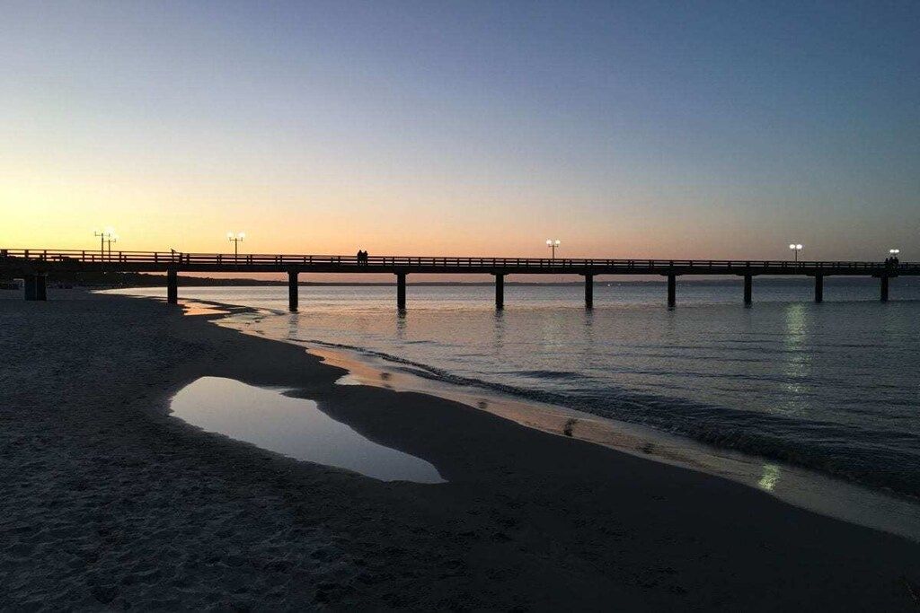 A beach with a pier extending into calm waters, a clear sky, and a few people walking on the pier.