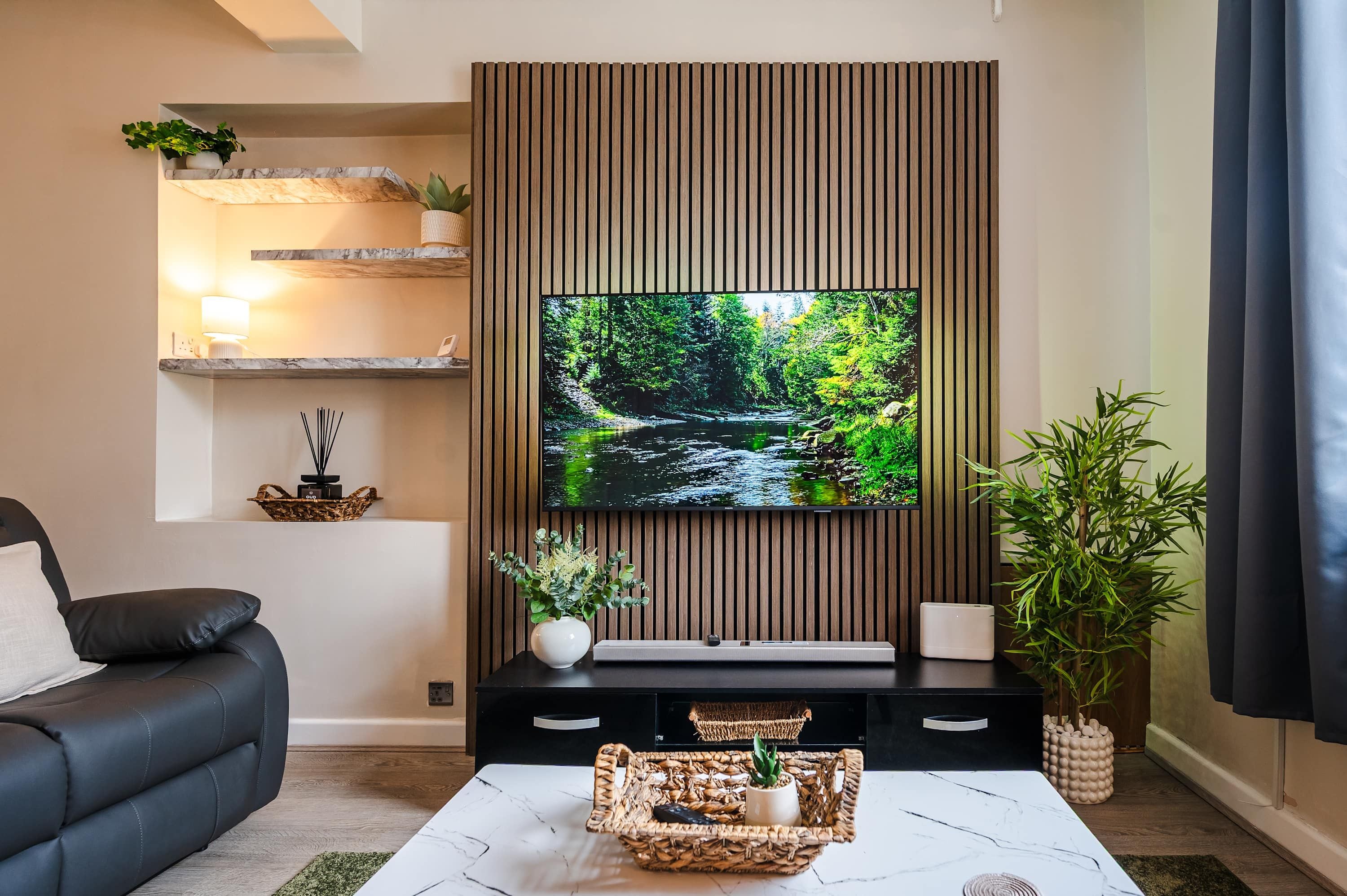 A modern living room with a flat-screen TV mounted on a wooden panel wall, a black sofa, a white coffee table with decorative items, and a potted plant.