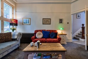 A living room with a red sofa, a coffee table, and a lamp. There are framed pictures on the wall and a staircase in the background. - Finest Retreats | Hall Bank (Buxton)