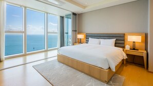 Habitación de hotel moderna con una cama grande, dos mesitas de noche con lámparas, vistas al mar y un ventanal amplio.