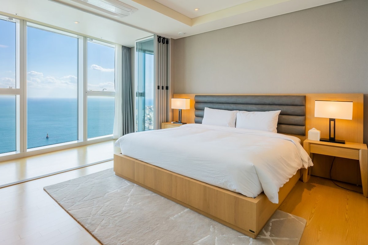 A modern hotel room with a large bed, two bedside tables with lamps, a view of the sea, and a large window.