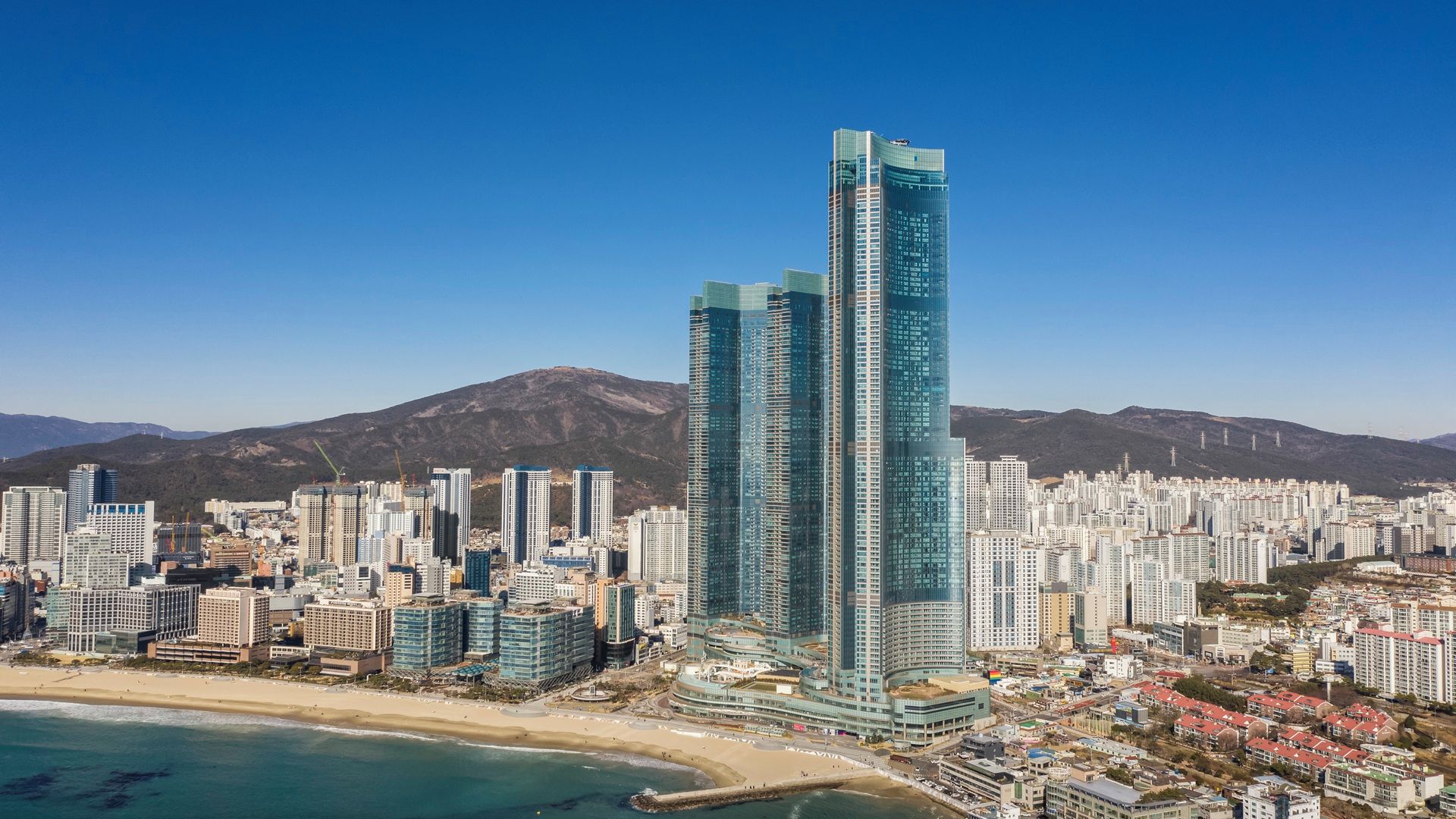 Un paisaje urbano costero con rascacielos, una playa y montañas al fondo.