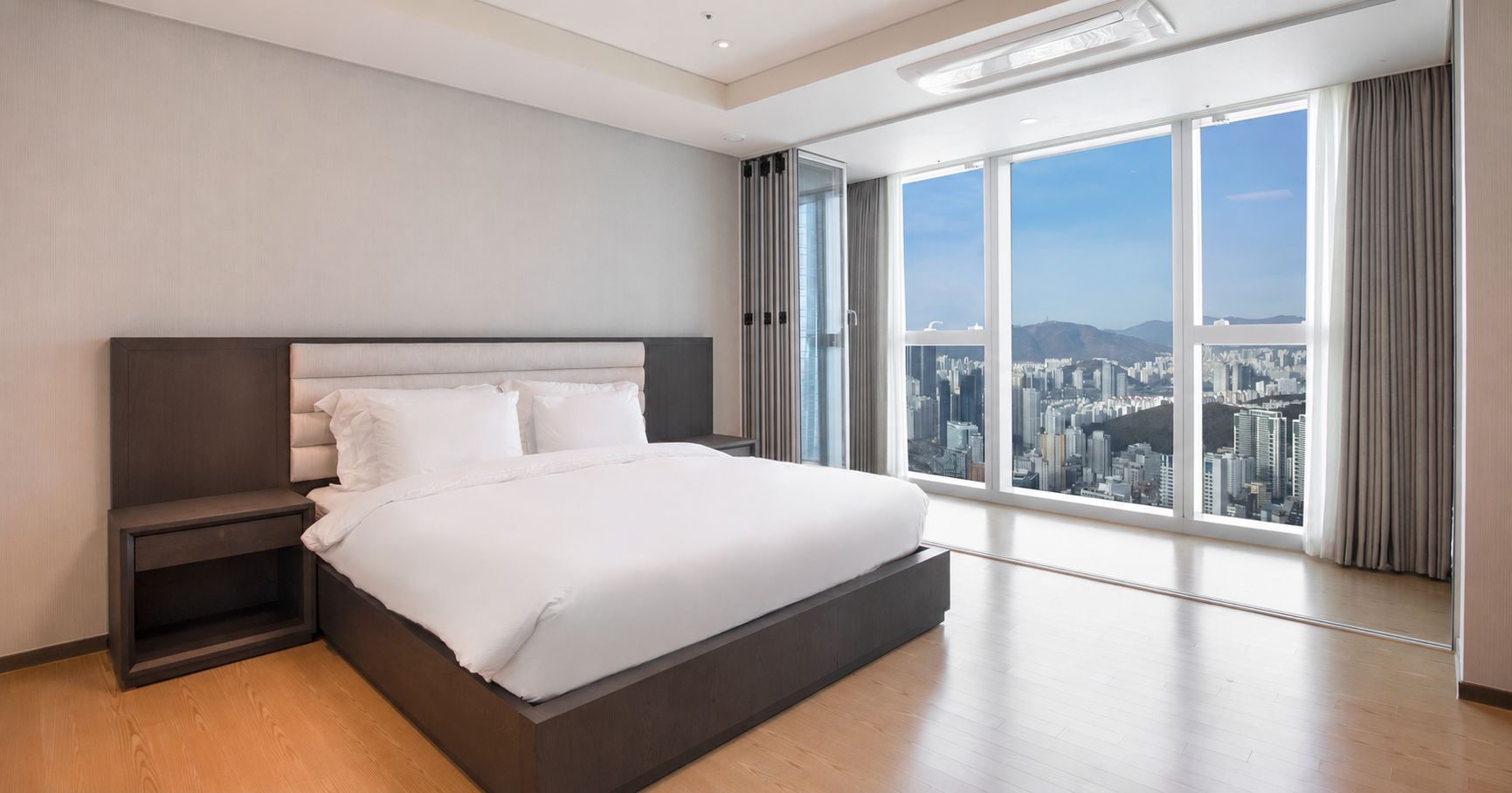 Un dormitorio moderno con una cama grande, una mesita de noche y vistas a la ciudad a través de amplias ventanas.