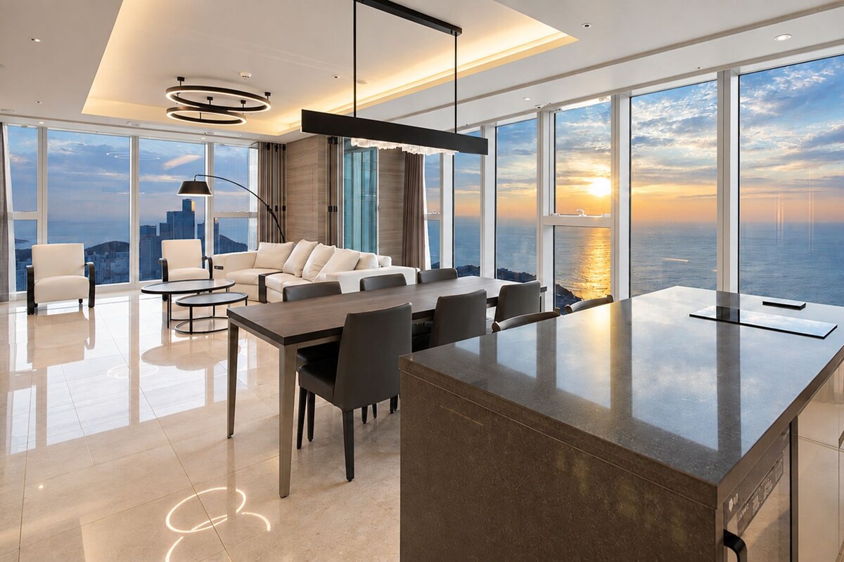 A modern interior with a dining area, sofas, and a large window offering a view of the sea and cityscape.