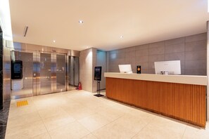 A modern reception area with a wooden reception desk, glass elevator doors, and a digital information kiosk. - LCT Residence the Drayton (Busan)
