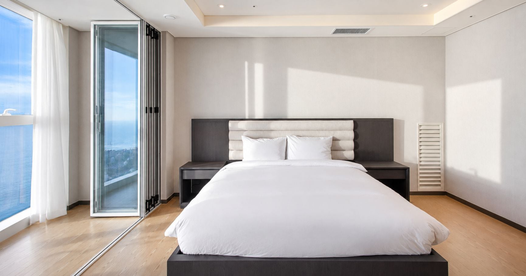 A modern bedroom with a large bed, a nightstand, a window with curtains, and a view of the city.