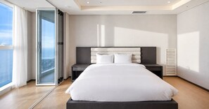 A modern bedroom with a large bed, a nightstand, a window with curtains, and a view of the city. - LCT Residence the Drayton (Busan)