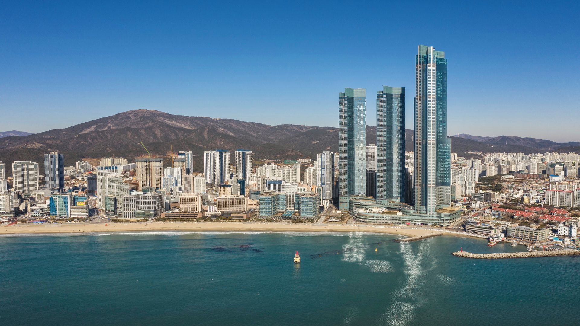 A coastal cityscape with high-rise buildings, a beach, and mountains in the background.