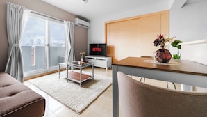 A modern living room with a glass coffee table, a sofa, a dining table with chairs, a TV, and a city view through large windows. - E-US Naebo 403 (Sapporo)