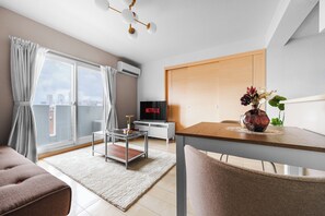 En moderne stue med et glasbord, en sofa, et spisebord med stole, et fjernsyn og udsigt over byen gennem store vinduer.